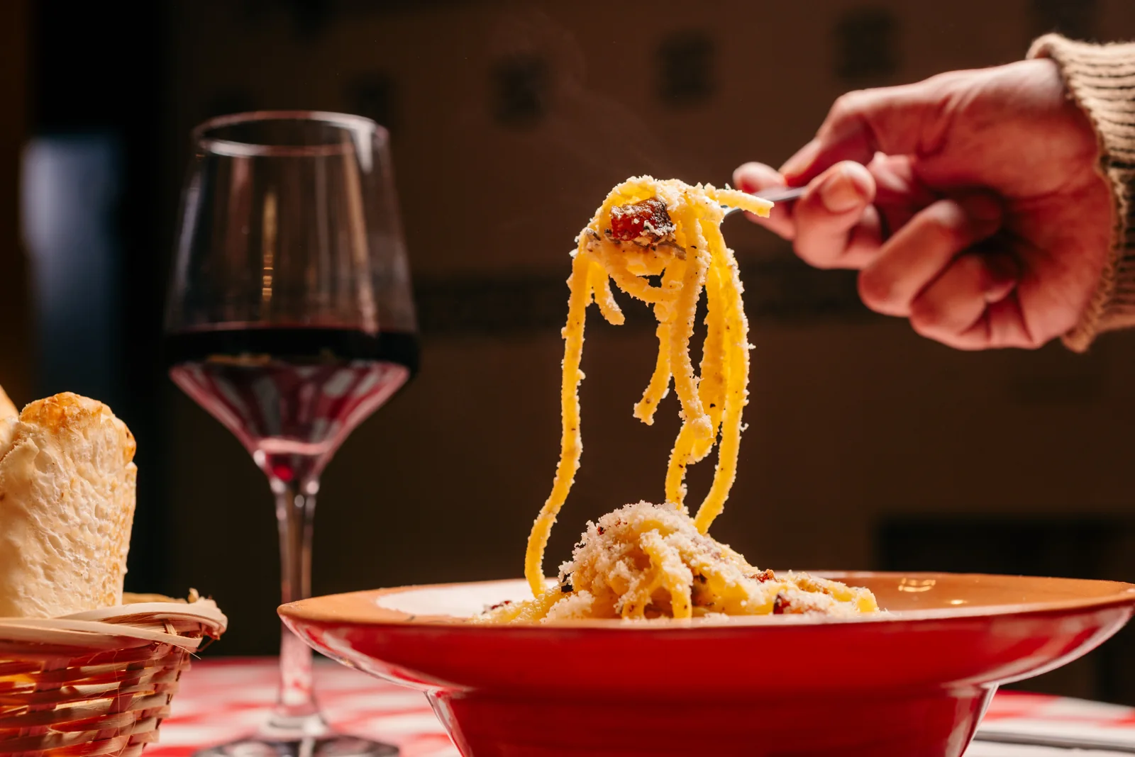 Un tenedor levantando pasta sobre un plato de La Piccola, junto a una copa de vino